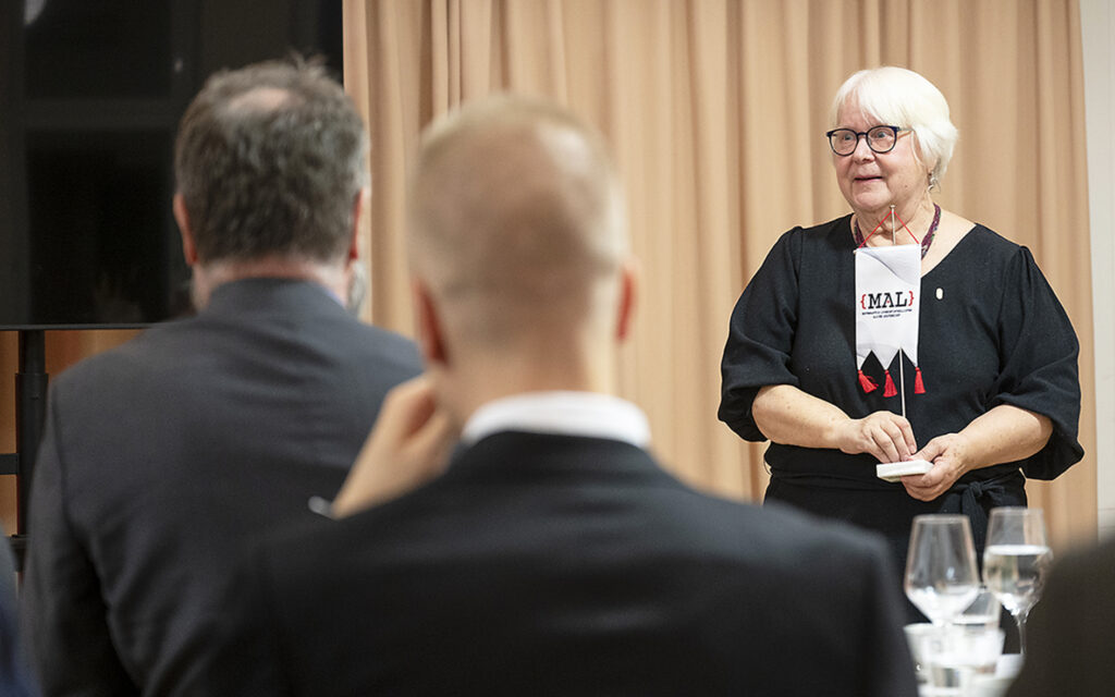 Vaaleahiuksinen nainen seisoo MAL-viiri kädessä. Kuvan etualalla kaksi miestä selin kameraan.