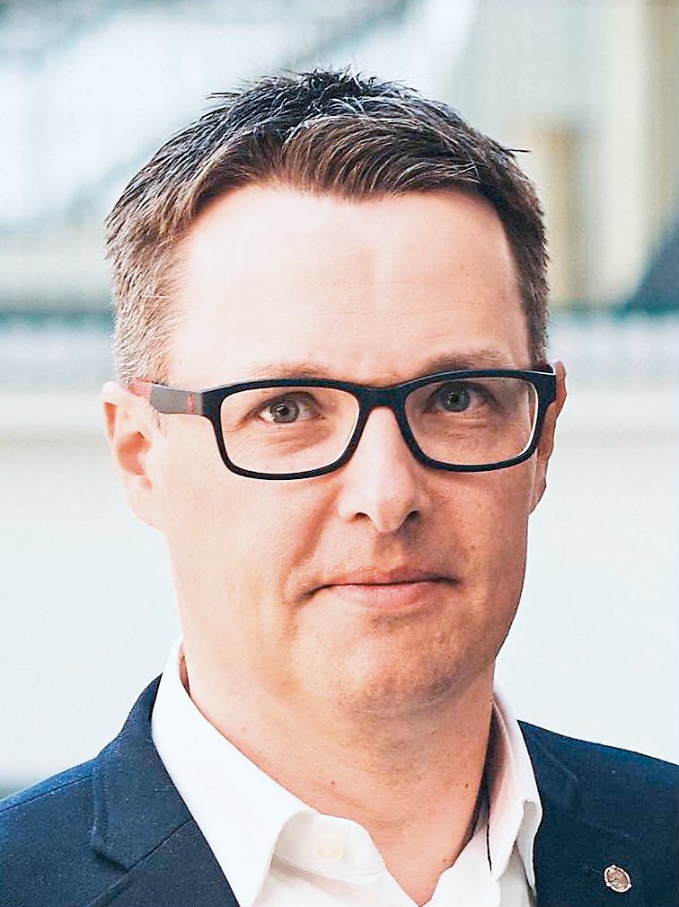 Lyhythiuksinen ja silmälasipäinen henkilö käyttää tummaa bleiseriä valkoisen kauluspaidan päällä ja katsoo suoraan kameraan neutraalilla ilmeellä.